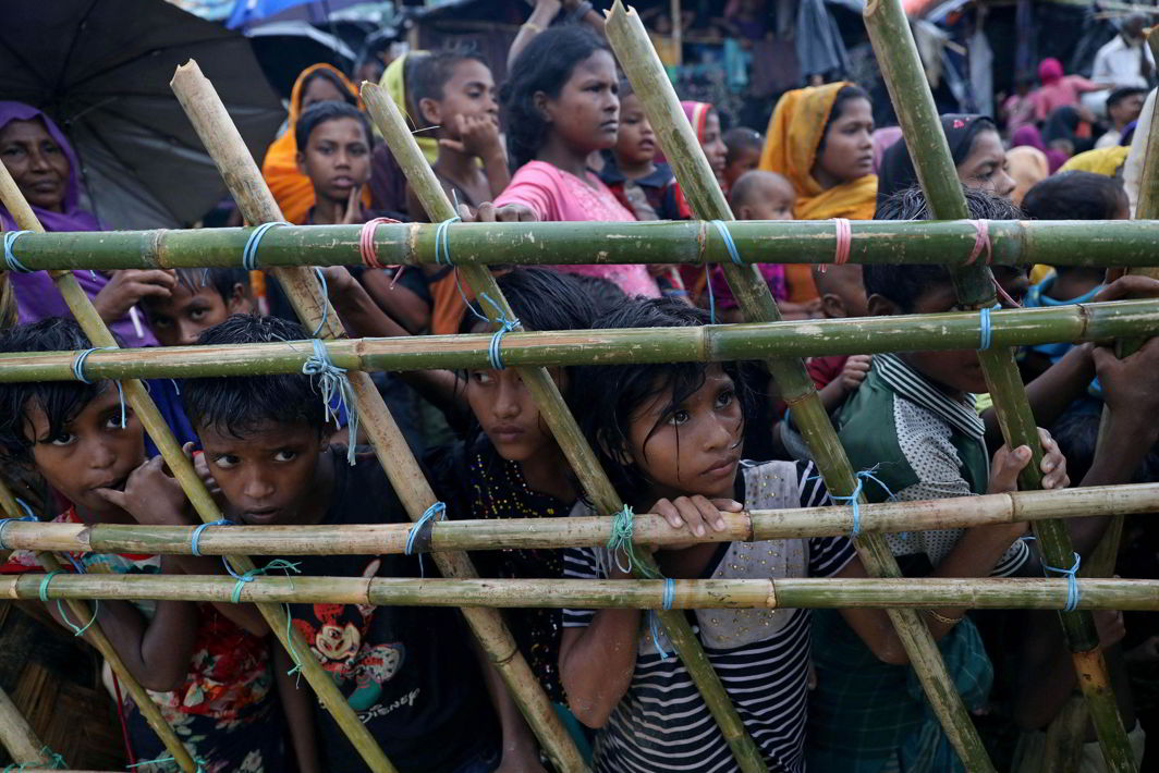 Rohingya refugees look through a fence as they wait outside of aid distribution premises at a refugee camp. Photo: UNI
